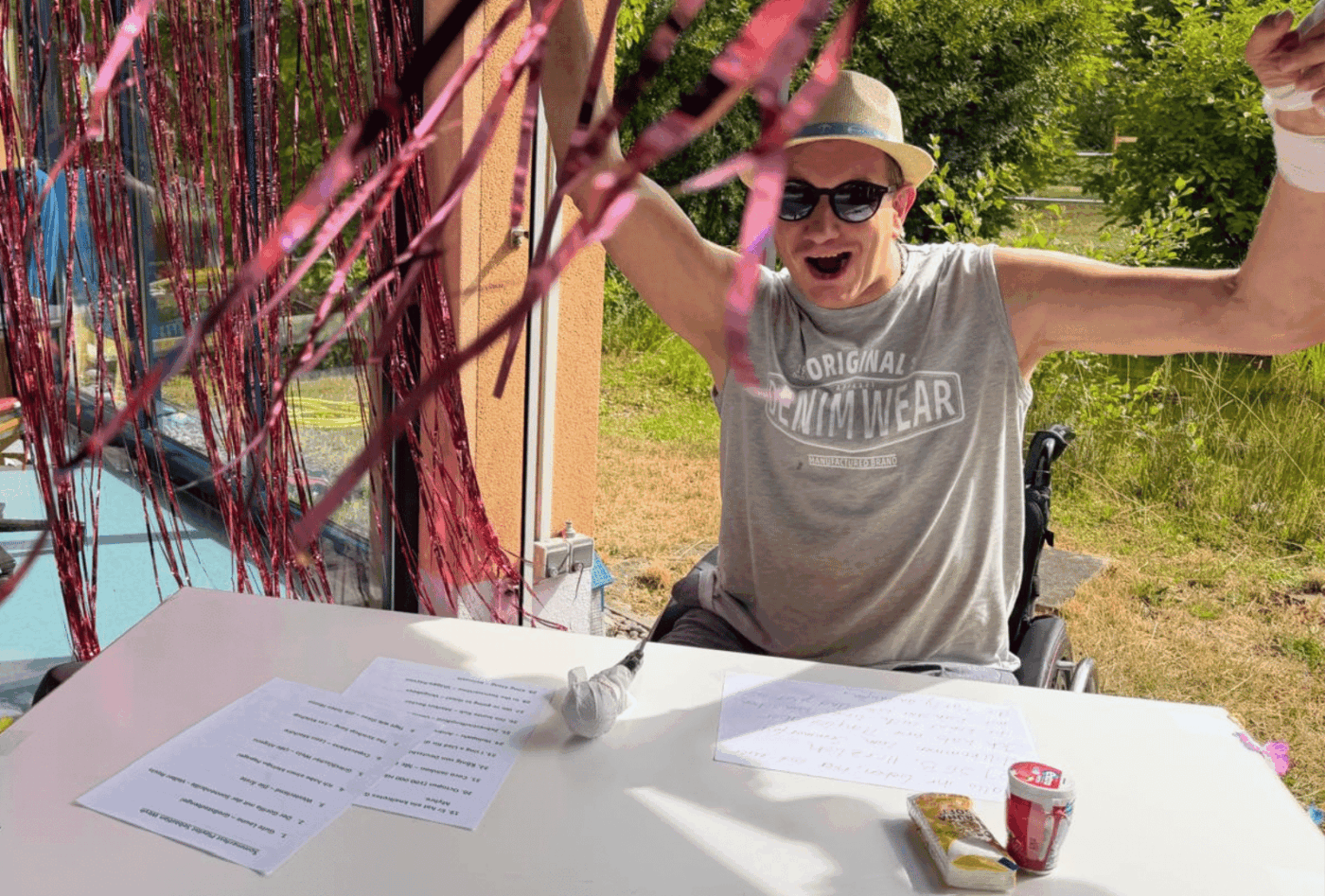 Ein Klient des Förderbereichs legt beim Sommerfest als DJ Musik auf. Im Rollstuhl sitzend hebt er lachend beide Arme und trägt Sonnenhut und Sonnenbrille.