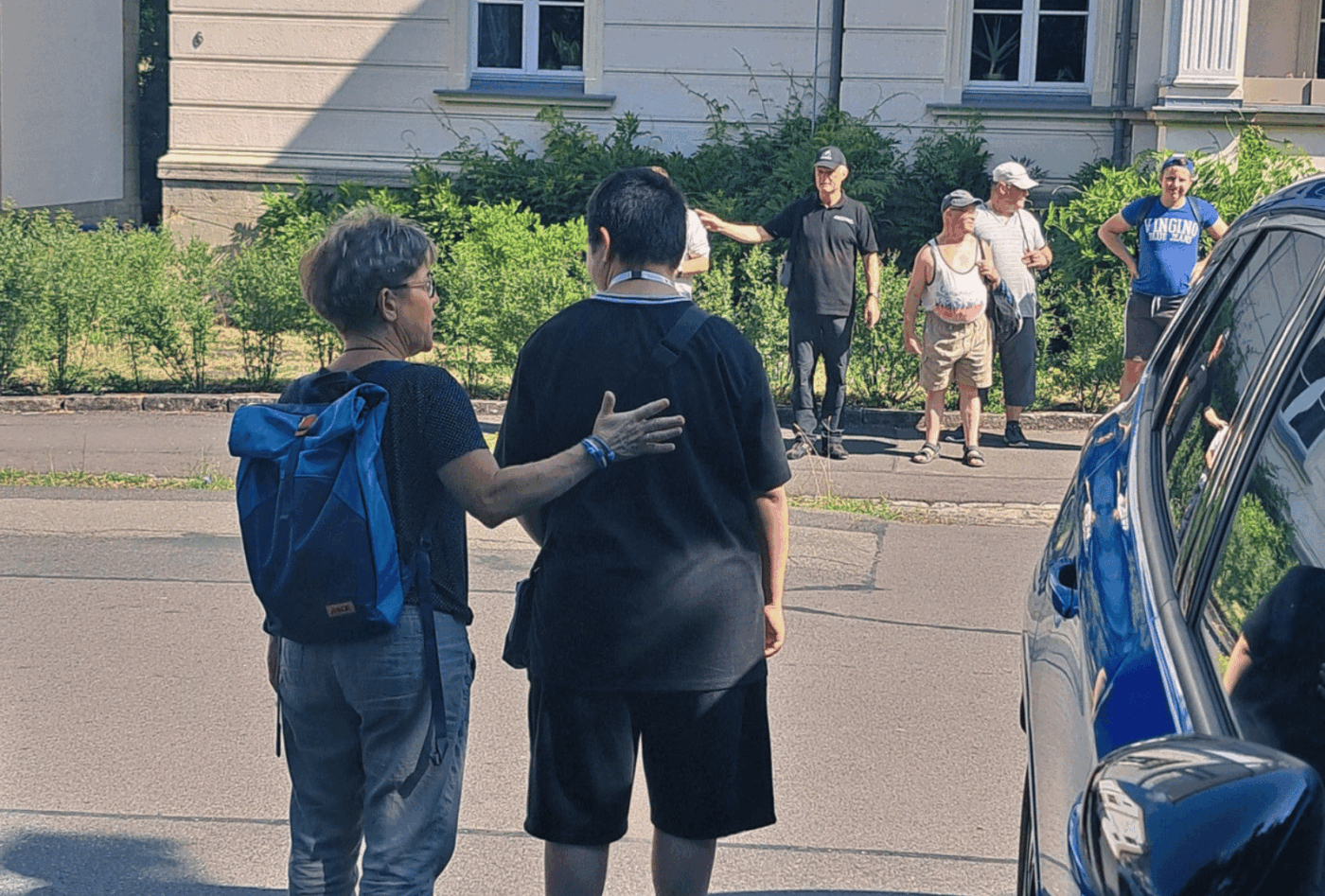 Ein Beschäftigter übt mit einer Mitarbeiterin des Sozialteams das sichere Überqueren einer Straße. Auf der anderen Straßenseite wartet die Gruppe mit einem Polizeibeamten.